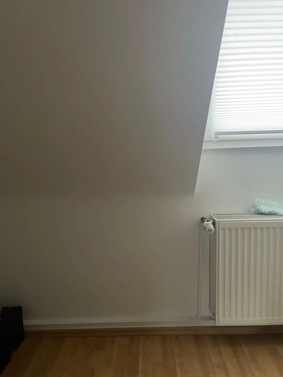 Weiße Dachschrägenwand mit Fenster und Jalousie, daneben ein weißer Heizkörper über einem Holzlaminatboden.
